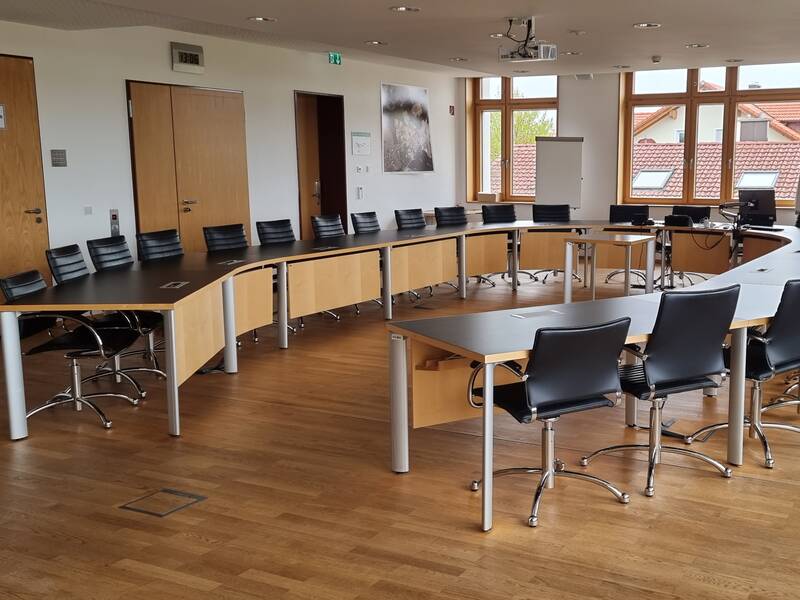 Ein Konferenzraum mit einem langen hufeisenförmigen Tisch, umgeben von vielen schwarzen Stühlen, ausgestattet mit moderner Technik und großen Fenstern.